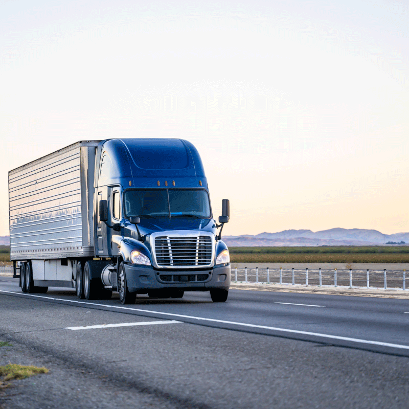 Blue semi-truck driving down a highway at sunset