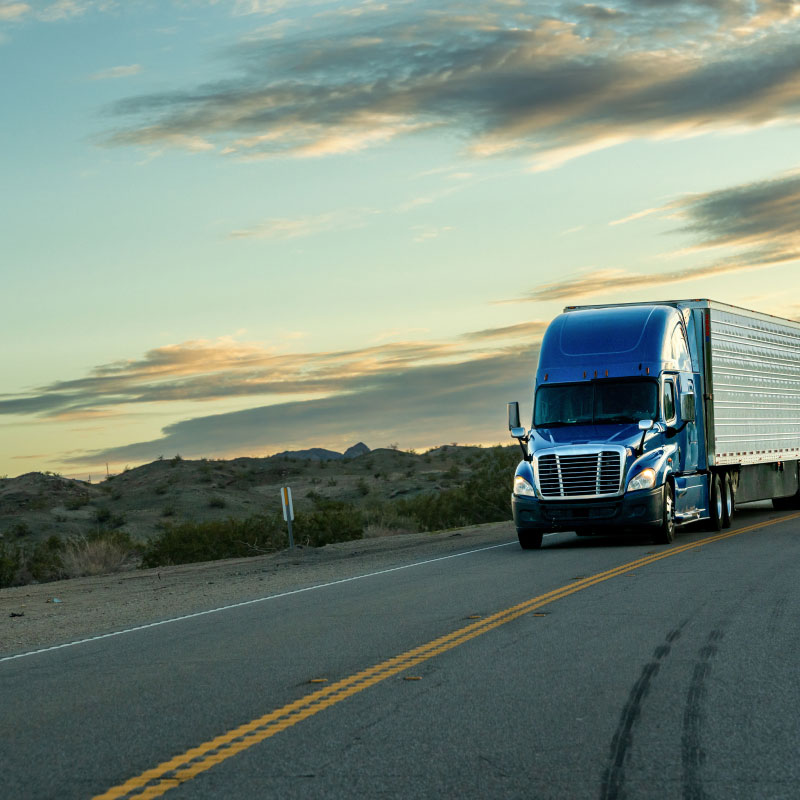 Blue semi truck driving down desert road during dusk.