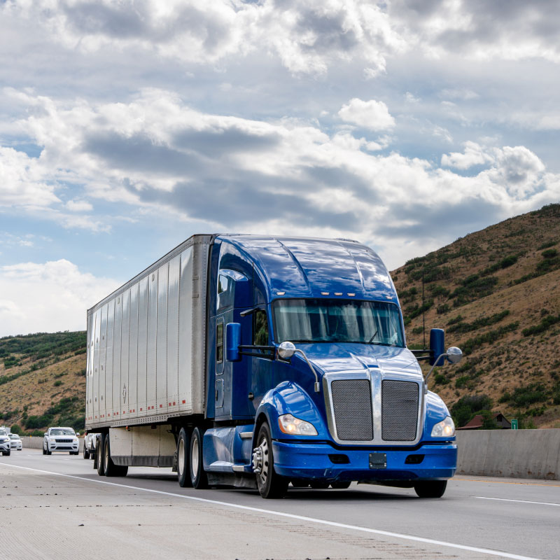 A blue semi truck driving down a road with evergreens in the background.