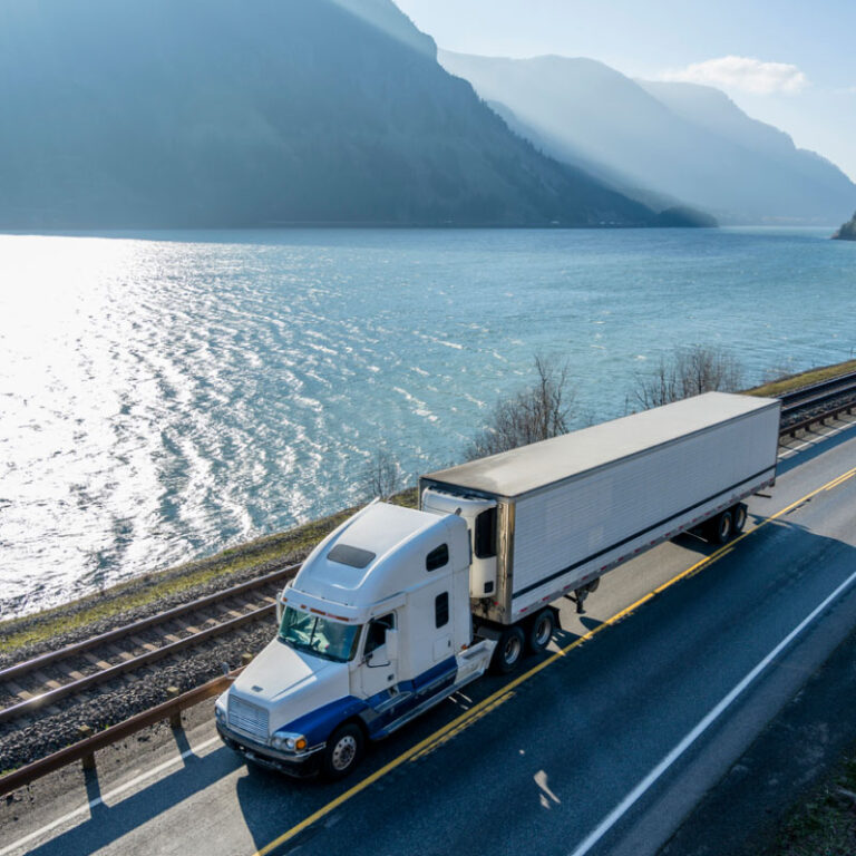 A temperature controlled semi truck driving on the road along a railroad and river in Columbia Gorge area mountain ranges.