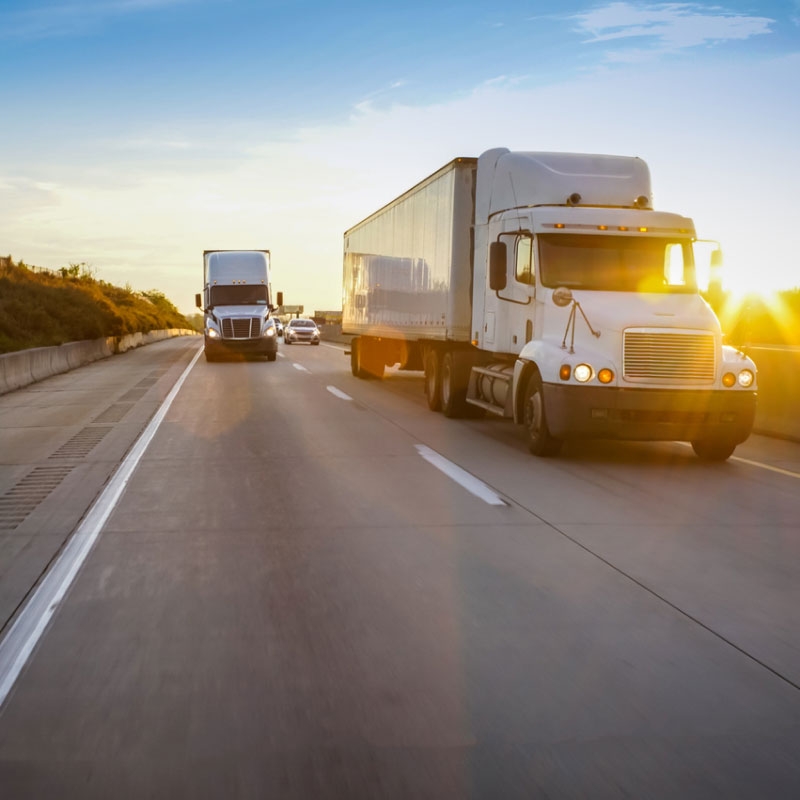 Two white semi trucks driving down the highway at sunset