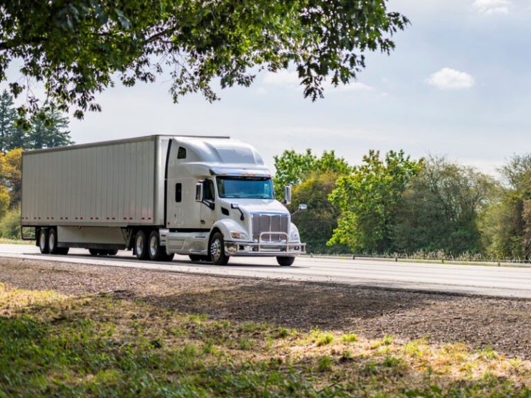 A white commercial semi truck with a dry van truck bed driving on highway to deliver cargo.