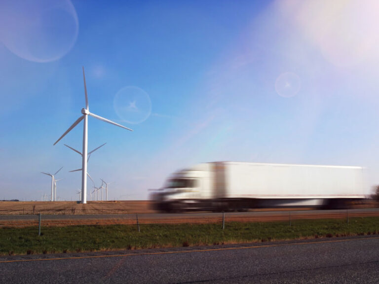 A blurred-motion white semi truck driving down past wind turbines on a road.