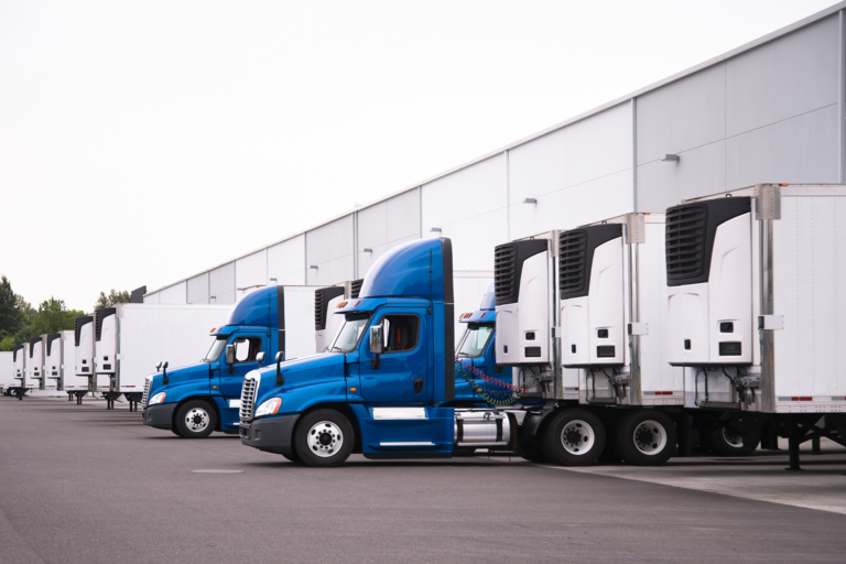 A line of blue semi-trucks with temp controlled truck beds parked at a warehouse loading dock.